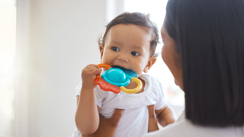 Mother holding up a baby who is chewing on a teething toy and looking off to the side