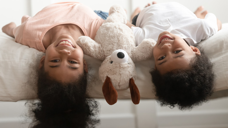 Two kids hanging upside down on a bed with a stuffed animal