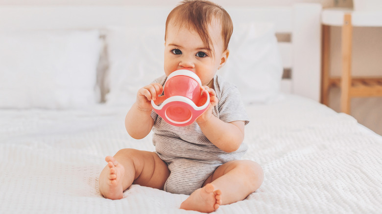 Baby sitting on bed drinking from sippy cup
