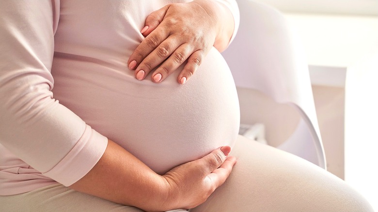 Pregnant woman wearing a long-sleeved pink shirt and tan pants, holding her belly with both hands