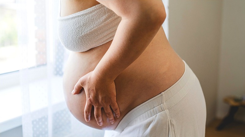 A woman in a white terrycloth bra leaning over and touching her pregnant belly with her left hand
