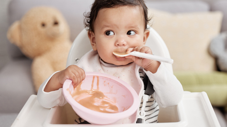 A baby looks off to the side while holding a spoon of pureed fruit to her mouth