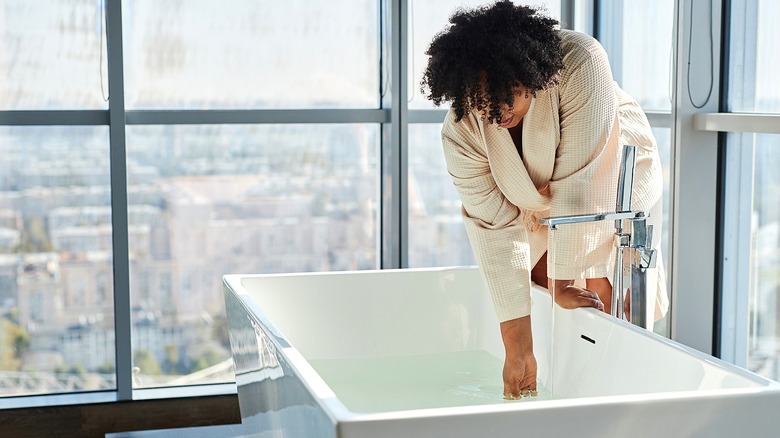 Black woman in a cream-colored robe leaning over a modern bathtub filling with water
