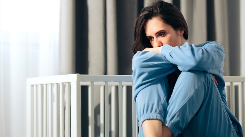 A mournful-looking woman in a blue sweat suit holding her knees in front of an empty white crib