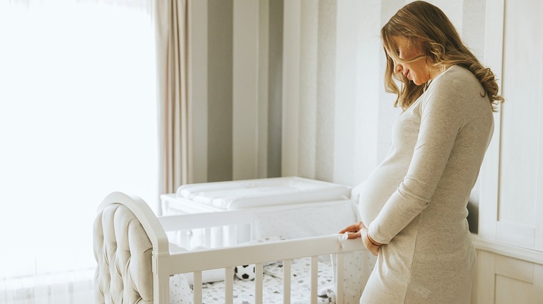 A pregnant woman holding her stomach while holding the side of a white, bassinet-style crib