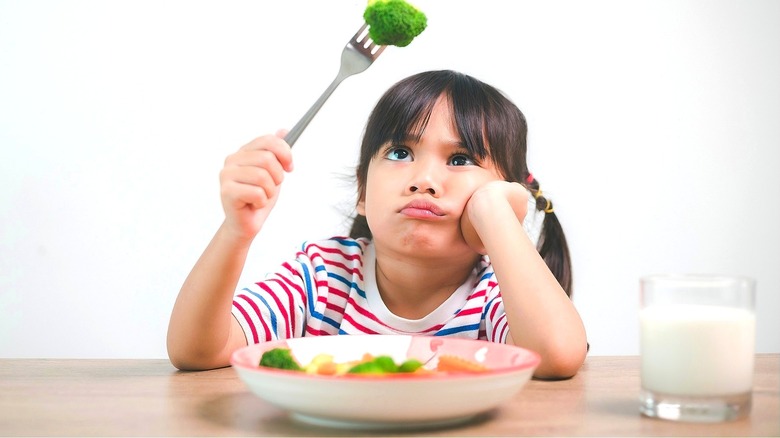 A child in pigtails sitting at a dining table, looking questionably at a piece of broccoli on a metal fork