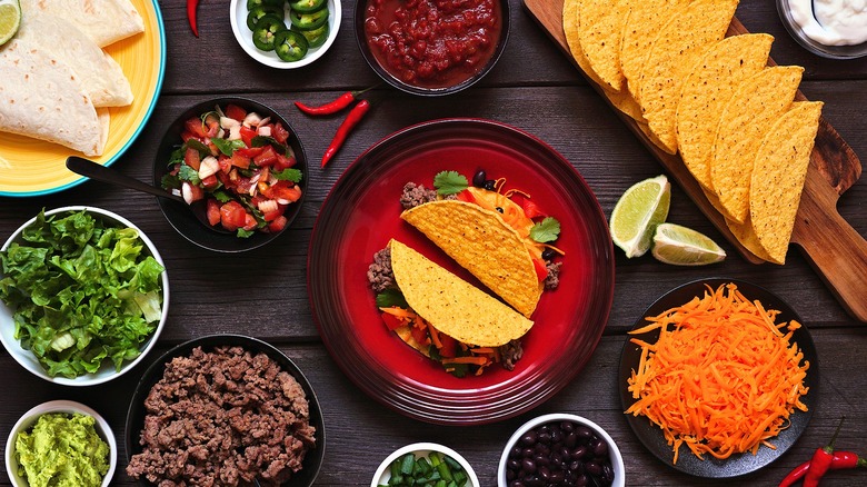 Crunchy tacos on a plate surrounded by various taco ingredients in small bowls like ground beef, lettuce, salsa, and black beans
