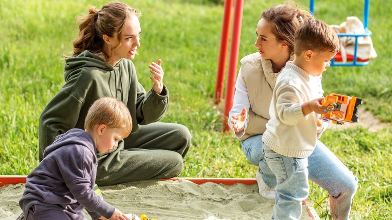 Two moms with their toddler sons sitting near a sandbox talking as their kids play