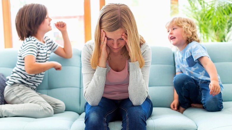 A woman holding her face in her hands while two young children sit and talk behind her