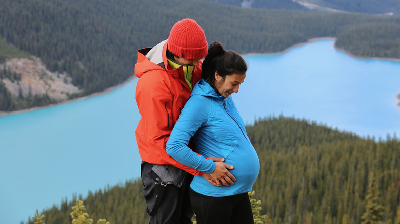 A man stands behind a pregnant woman with both their hands cupping the woman's pregnant belly while standing in front of a mountain and lake landscape
