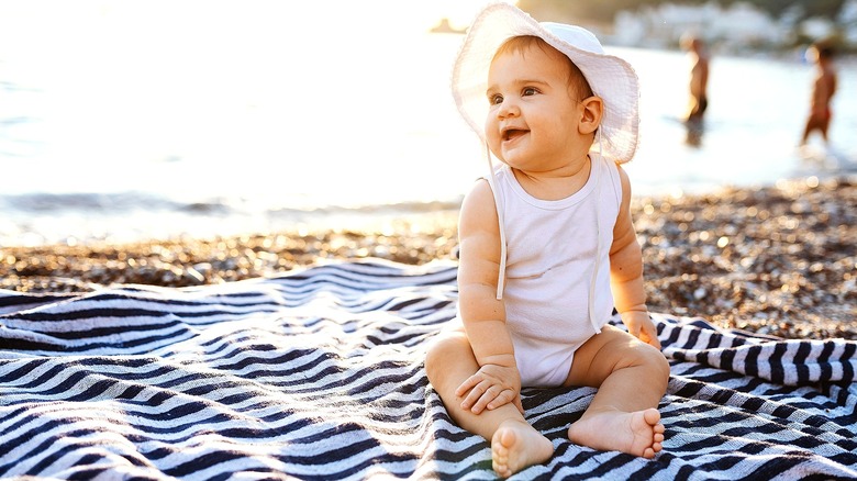 A baby in a white tank onesie and floppy white hat sitting on a black and white striped towel on the beach