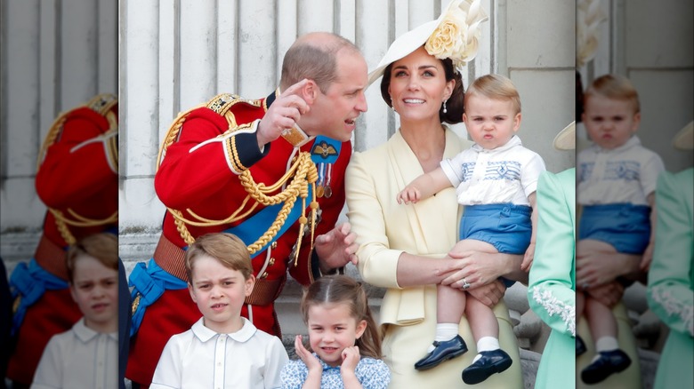 William, Prince of Wales talking to Prince Louis while he scrunches his nose while Catherine, Princess of Wales holds him and Prince George and Princess Charlotte watch from the royal balcony