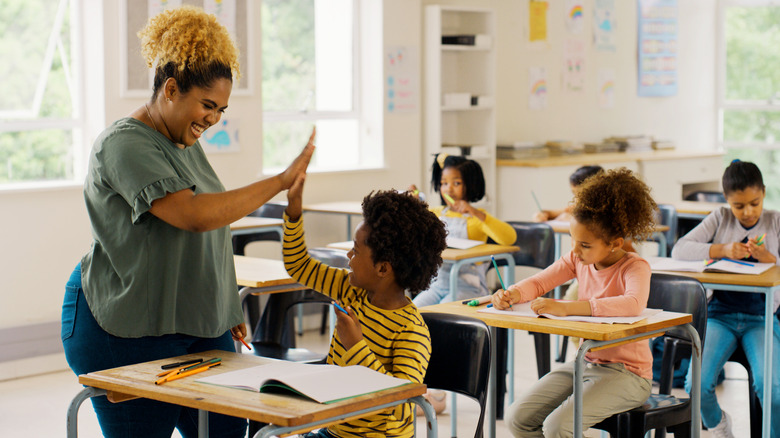 A teacher stands over a child seated at a desk and gives him a high-five while other students work in the background