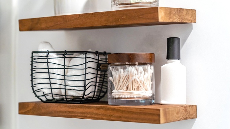Two floating wooden shelves holding a dark wire basket with towels and a small glass container with Q-Tips