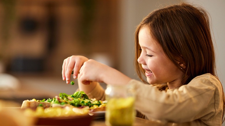 Smiling girl holding peas above a plate of bread and leafy greans