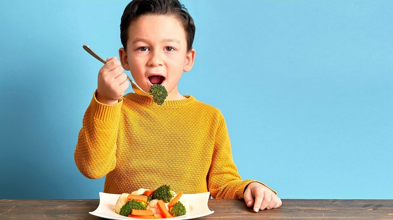 Small boy wearing a gold sweater holding a fork of cooked broccoli up to his mouth