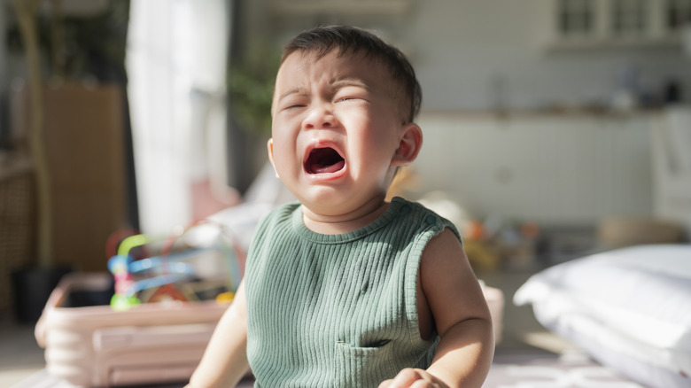 A baby with an emotional expression sits upright with his mouth open in the middle of crying