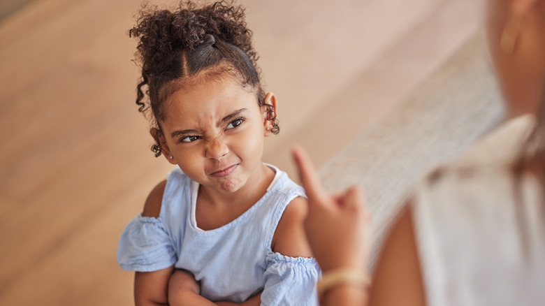 A little girl looks up at an adult defiantly with crossed arms and a scrunched facial expression