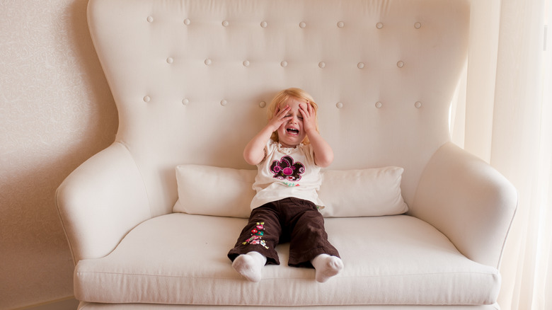 Child with gripping their face with their hands with an emotional expression while sitting in a large white chair