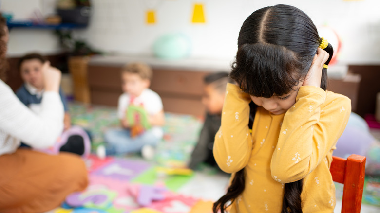 A girl sits with her back to a teacher and a group of other students with her hands up to her ears, as if refusing to listen and participate