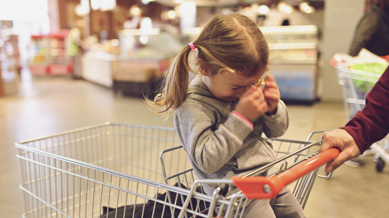 A little girl sits in a shopping cart with her hands to her face making an upset gesture
