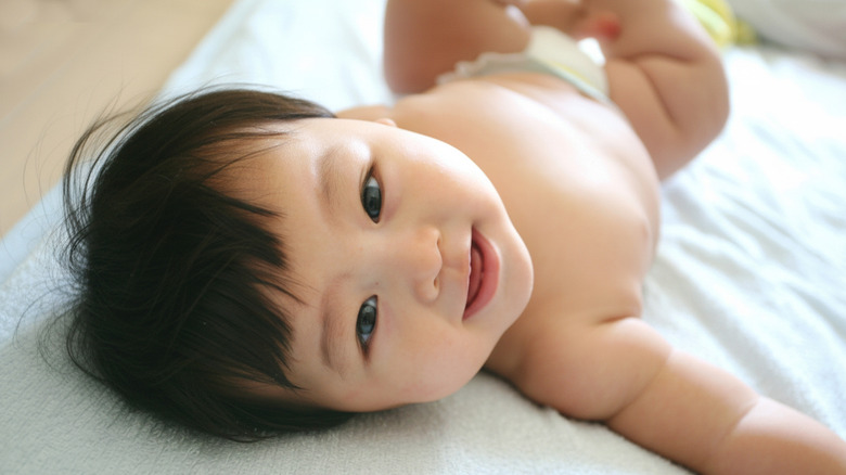 A baby lies on a bed looking behind him smiling at the camera