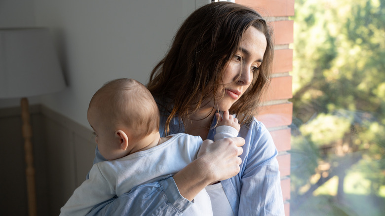A lonely mother stares sadly out a window holding her baby