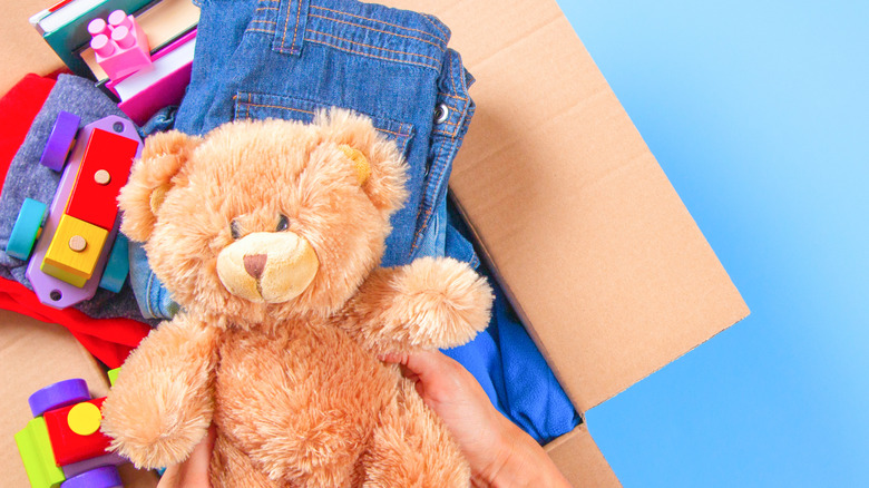 Top view of a person's hands reaching into a box of children's donations with a teddy bear, clothing, books, and toys