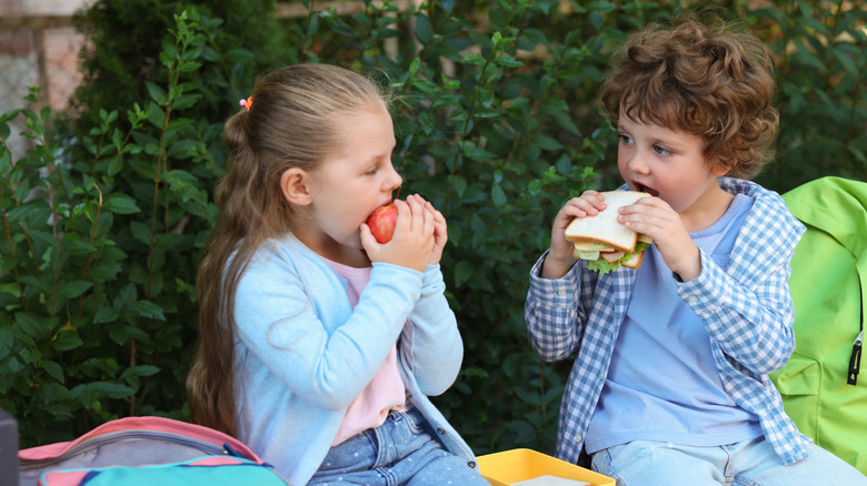 A young girl sits on a bench outside eating an apple and facing a young boy, who holds a sandwich up to his face