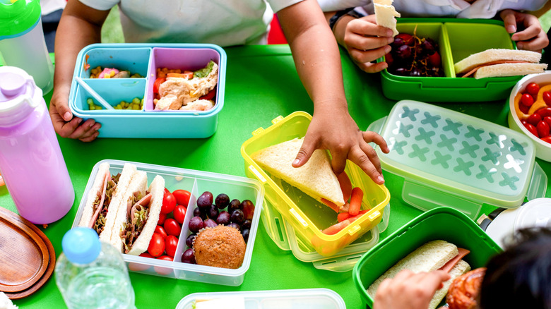 Children sit around a bright green table reaching their hands into various plastic containers with small portions of food