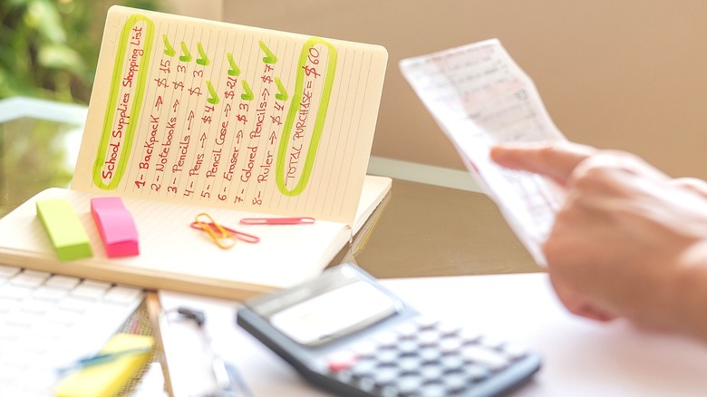 A person holding and pointing to a receipt over a desk with a calculator and school supply shopping list