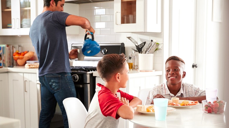 Two young boys sitting at a round dining table while a man pours hot water into a mug in the background