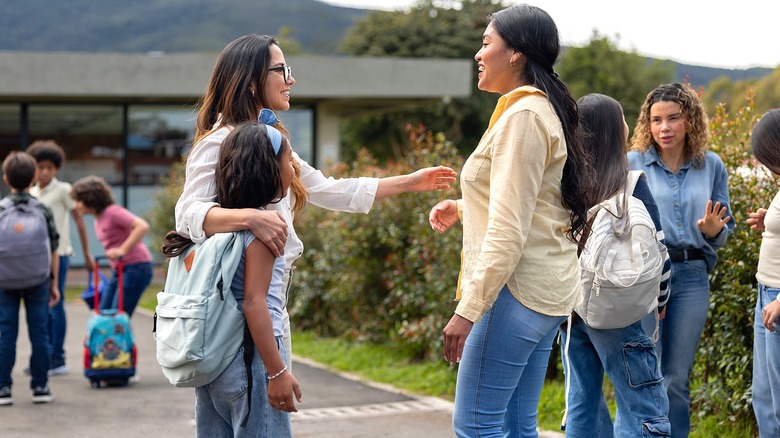 Two women with long, dark hair talking to one another outside of a school building surrounded by students in backpacks