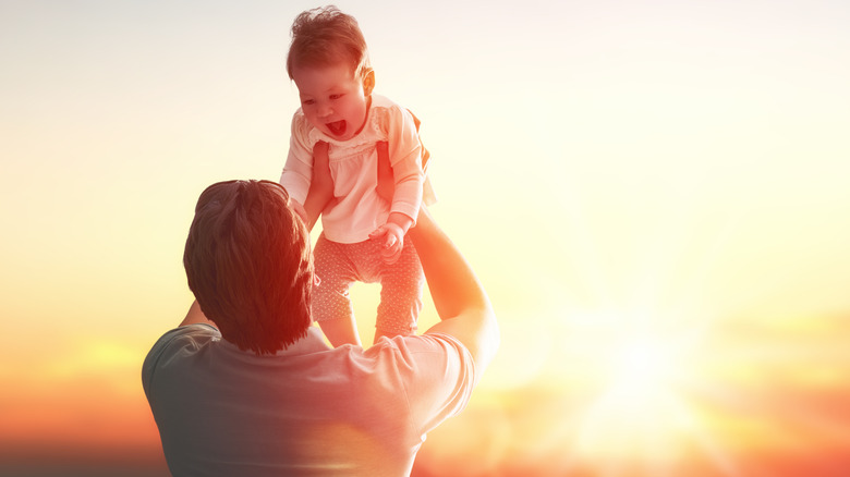 father holding baby high in the air while the sun is behind her