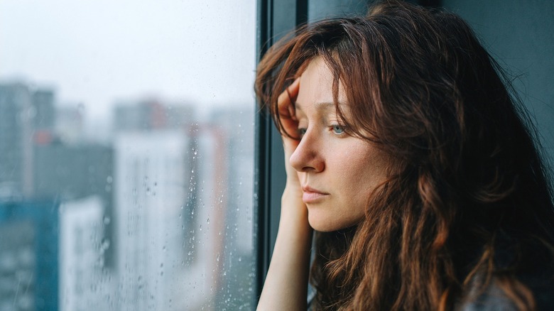 A distressed woman with long reddish-brown hair looking out a rainy window in a city