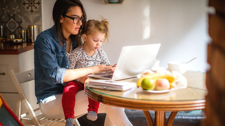 A woman with glasses holding a small child on her lap while typing on a laptop computer