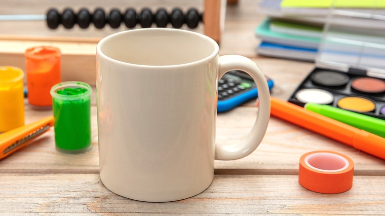 White coffee mug with handle surrounded by open pots of paint, paint palette, and colorful pens on a wooden surface
