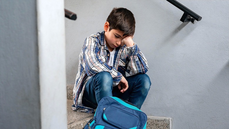 A sad-looking boy with short, dark brown hair holding his face while sitting on steps with a blue backpack at his feet