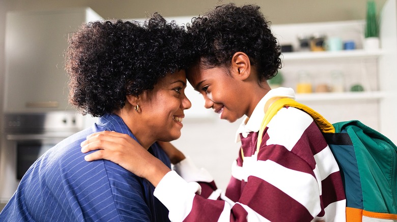 A mom and her young son wearing a backpack, forehead to forehead smiling at each other