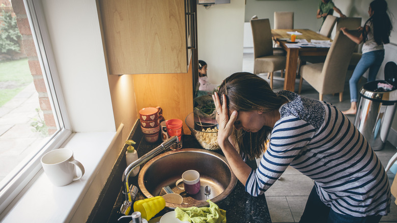 A stressed-out mom rests her head in her hands in front of a sink full of dishes with her kids running in the background