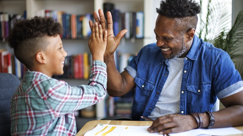 An African American father and son high-fiving each other while sitting together at a table over an open workbook
