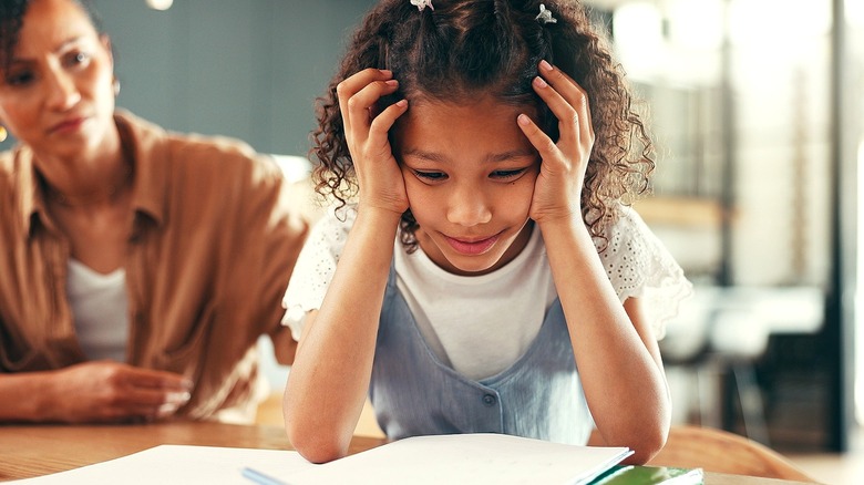 Child with dark curly hair holding her face over a book with her mother looking concerned in the background