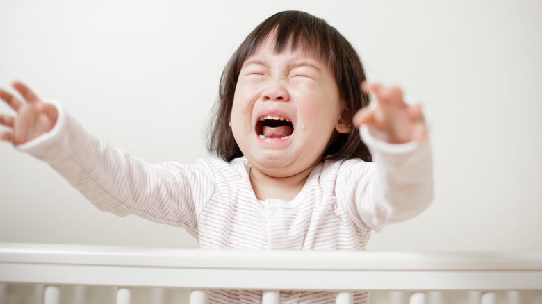 Toddler girl stands in crib crying reaching out her hands