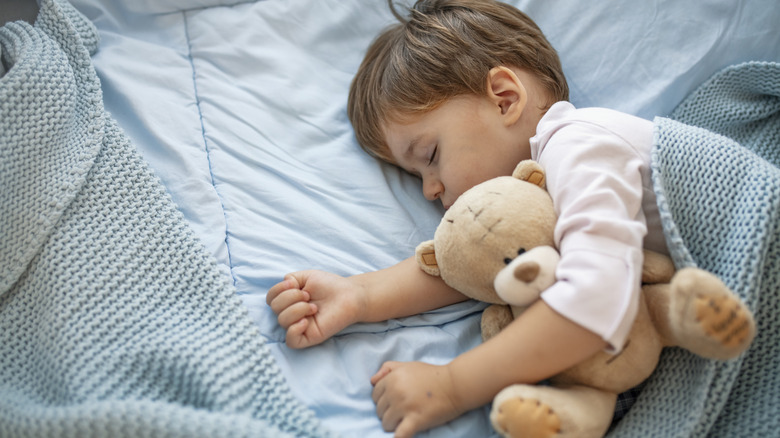 Baby tucked in with blue sheets, a brown stuffed bear, and a blue woven blanket