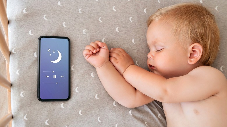 Top-down view of a baby napping in a crib next to a smartphone playing sleep sounds