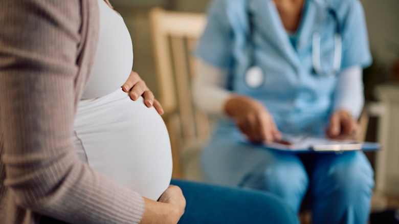 Woman sitting in a chair holding her pregnant stomach with a doctor holding a clip board in the background