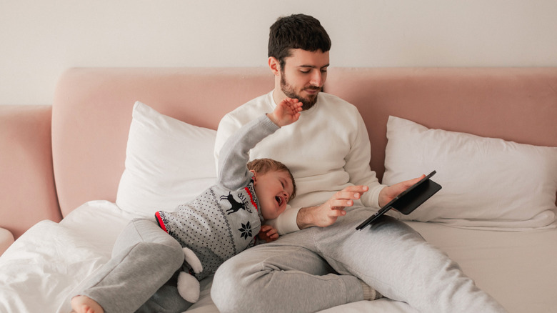 A father holds a tablet on his lap in bed while a little boy cries and flails his arms next to him