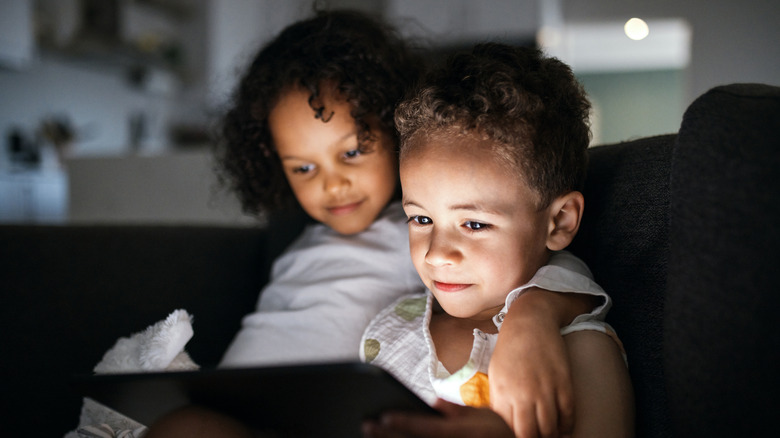 A young boy and girl sit together on a couch staring at a lit tablet screen